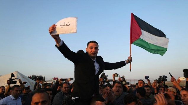 يرفع فلسطينيون عريس وهو يلوح بعلم فلسطيني ويحمل لافتة باسم قرية عائلته في إسرائيل على حدود غزة مع إسرائيل (خلفية)، شرق جباليا، في 29 مارس 2018، قبل معسكر الاحتجاج. ومدته ستة أسابيع. (AFP PHOTO / MAHMUD HAMS)