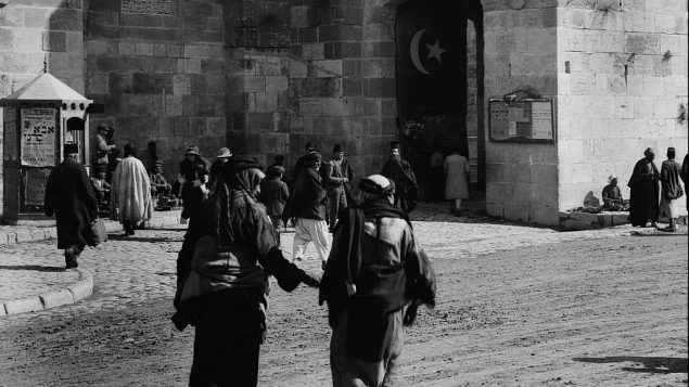 الموقع المحدد غير معروف، رجلان يسيران باتجاه البلدة القديمة، والعلم التركي ظاهر عند الباب. (Unknown)