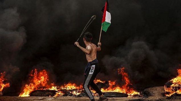 متظاهر يحمل العلم الفلسطيني بينما يستخدم مقلاع لرشق الحجارة باتجاه جنود اسرائيلبيين خلال اشتباكات بالقرب من الحدود مع اسرائيل، شرقي مدينة غزة، 26 اكتوبر 2018 (Mahmud Hams/AFP)