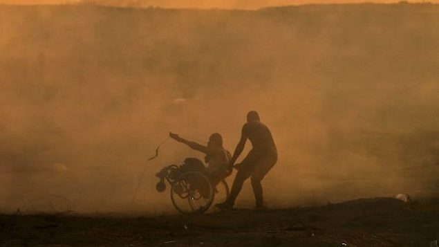 فلسطيني يسحب فلسطينيًا آخر على كرسي متحرك وسط سحابة الغاز المسيل للدموع الذي أطلقته القوات الإسرائيلية خلال اشتباكات شرق مدينة غزة بالقرب من الحدود الإسرائيلية في 2 نوفمبر 2018. (Mahmud Hams/AFP)