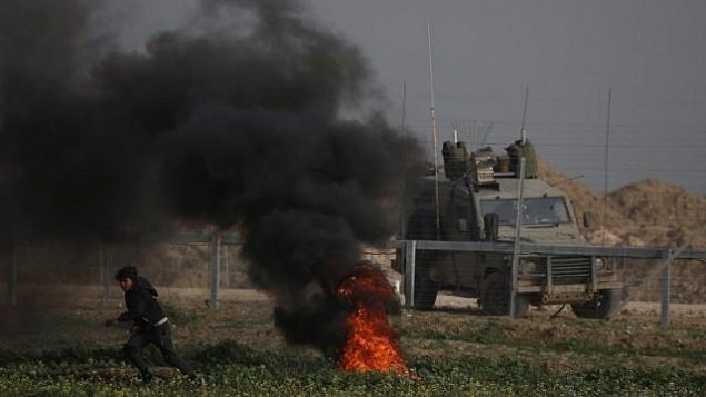 أحد المتظاهرين الفلسطينيين يركض بالقرب من الإطارات المحترقة خلال مظاهرة بالقرب من السياج على طول الحدود مع إسرائيل، شرق مدينة غزة، في 15 فبراير، 2019. تظهر سيارة عسكرية إسرائيلية على الجانب الآخر من السياج. (Said Khatib/AFP)