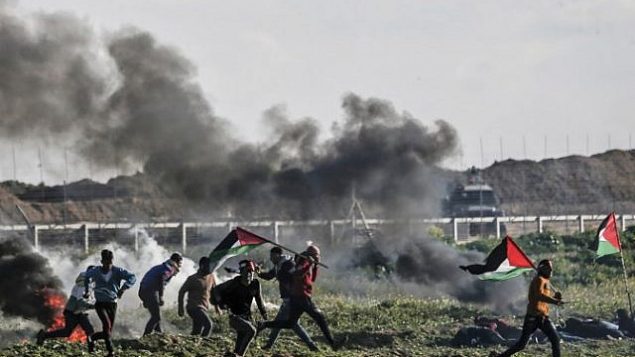فلسطينيون يحملون الأعلام الفلسطينية يمرون من أمام إطارات محترقة خلال مواجهات بالقرب من السياج الحدودي مع إسرائيل، شرق مدينة غزة، 22 فبراير، 2019. (Mahmud Hams/AFP)