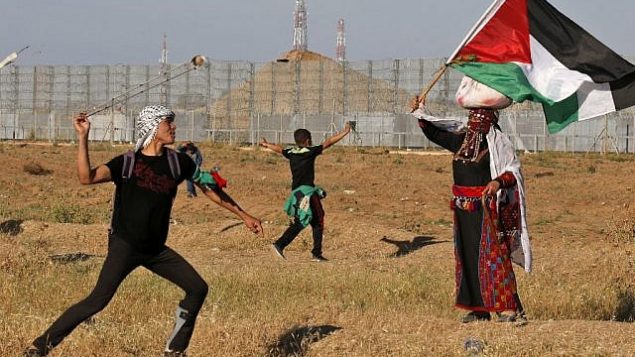 متظاهر فلسطيني يرشق القوات الإسرائيلية بحجارة وامراة ترتدي زيا فلسطينيا تقليديا تلوح بالعلم الفلسطيني، خلال تظاهرة بالقرب من الحدود مع إسرائيل، شرق مدينة غزة، 10 مايو، 2019. (Said KHATIB / AFP)