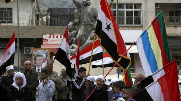 توضيحية: سكان هضبة الجولان يلوحون بالأعلام السورية ويقفون أمام صورة للرئيس السوري بشار الأسد خلال تظاهرة ضد اعتراف الرئيس الأمريكي دونالد ترامب بالسيادة الإسرائيلية على المنطقة المتنازع عليها، في بلدة مجدل شمس، 23 مارس، 2019.  (Jalaa Marey/AFP)