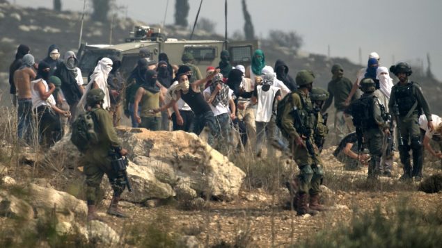 صورة توضيحية: جنود إسرائيليون يقفون امام مستوطنين إسرائيليين ملثمين يقومون بإلقاء الحجارة على متظاهرين فلسطينيين، خلال مظاهرة ضد البناء في بؤرة استياطنية إسرائيلية بالقرب من قرية ترمسعيّا الفلسطينية ومستوطنة شيلو، شمال رام الله في الضفة الغربية ، 17 أكتوبر 2019 (JAAFAR ASHTIYEH / AFP)
