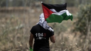شاب فلسطيني يحمل العلم الفلسطيني في الوقت الذي يقوم فيه الجيش الإسرائيلي بالبحث عن أنفاق تهريب على الحدود الشرقية لمدينة غزة، 15 مايو، 2016، في الذكرى ال68 ل’النكبة’، (AFP/Mahmud Hams)