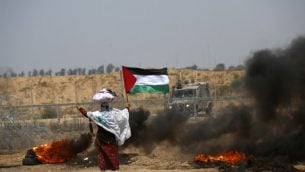 امرأة فلسطينية ترفع العلم الفلسطيني امام السياج الحودي مع اسرائيل، الى جانب إطارات مشتعلة، شرقي خان يونس، 8 يونيو 2018 (AFP/ SAID KHATIB)