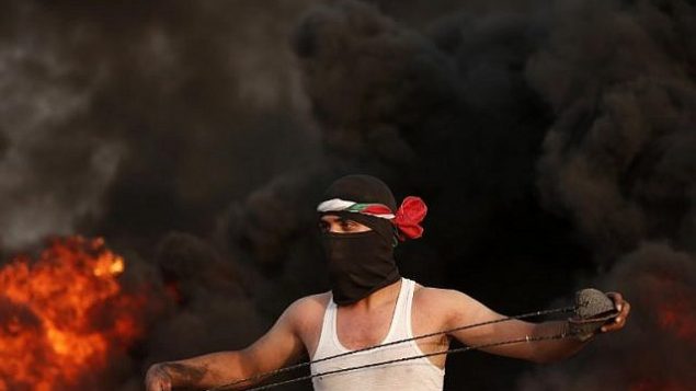 محتج فلسطيني ملثِّم يحمل مقلاعا ويقف أمام الدخان المتصاعد من الإطارات المحترقة خلال احتجاج بالقرب من الحدود مع إسرائيل شرق مدينة غزة في 31 آب / أغسطس، 2018. (AFP PHOTO / MAHMUD HAMS)