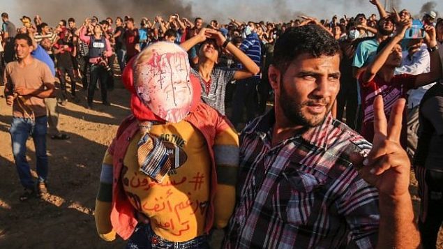 أحد المتظاهرين الفلسطينيين يلمح ببادرة النصر بينما يمسك دمية ترتدي قناعًا يصور وجه الرئيس الأمريكي دونالد ترامب مغطى بالدماء وقميصًا يحمل شعارًا عربيًا "مستمرون رغم أنف ترامب" ، أثناء مظاهرة على الحدود بين إسرائيل وغزة، شرق مدينة غزة في وسط القطاع في 7 سبتمبر 2018. (AFP/Said Khatib)