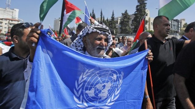 متظاهرون فلسطينيون يلوحون بعلم الأمم المتحدة والاعلام الفلسطينية امام بطاقة لاجئ ضخمة للرئيس الفلسطيني محمود عباس، خلال مظاهرة ضد قرار الولايات المتحدة في وقت سابق من العام وقف تمويل وكالة الامم المتحدة للفلسطينيين، في مدينة بيت لحم بالضفة الغربية، 26 سبتمبر 2018 (AFP/Musa Al SHAER)