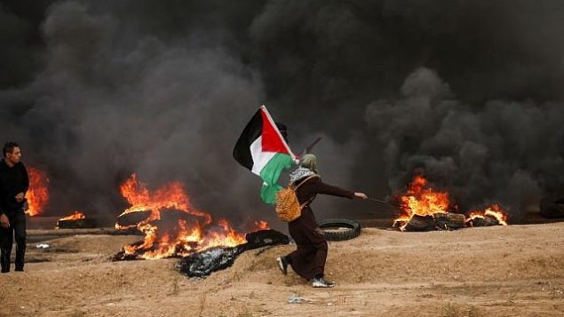 امرأة فلسطينية ترفع العلم الفلسطيني وسط دخان أسود من الإطارات التي أحرقها المتظاهرون، خلال اشتباكات قرب الحدود مع إسرائيل شرق مدينة غزة في 26 أكتوبر 2018. (MAHMUD HAMS / AFP)