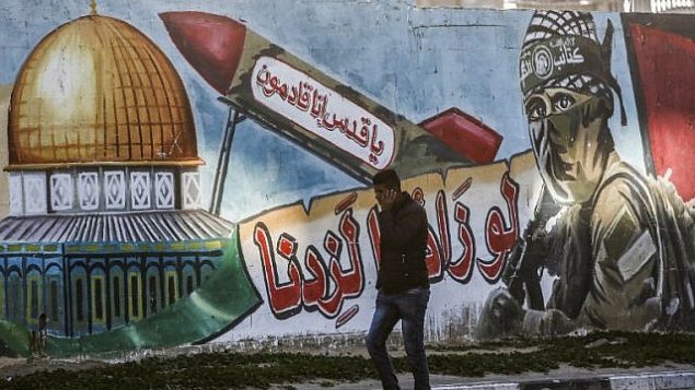 رجل فلسطيني يمر من أمام رسم لمقاتل ملثم من كتائب القسام، الجناح العسكري لحركة حماس، وإلى جانبه صاروخ تظهر عليه عبارة "يا قدس إنا قادمون" وقبة الصخرة، في أحد شوارع رفح في قطاع غزة، 14 مارس، 2019. (Photo by SAID KHATIB / AFP)