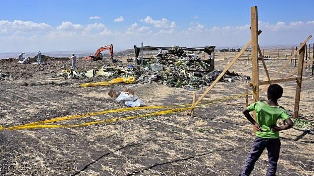طفل يتفرج على محققي الأدلة الجنائية وهم يقومون بتمشيط الأرض بحثا عن أدلة حمض نووي بالقرب من كومة من خطاب الطائرة في موقع تحطم طائرة الخطوط الجوية الإثيوبية من طراز ’بوينغ 737 ماكس 8’ ، 16 مارس، 2019، في قرية قريبة من بيشوفتو في منطقة أوروميا.  (TONY KARUMBA / AFP)
