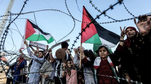 طفلة ترفع علم فلسطيني، بينما يرفع طفل اخر مفتاح خشبي يمثل حق العودة، خلف اسلاك شائكة عند الحدود بين قطاع غزة واسرائيل، شرقي مدينة غزة، 13 مايو 2018 (AFP/Mahmud Hams)