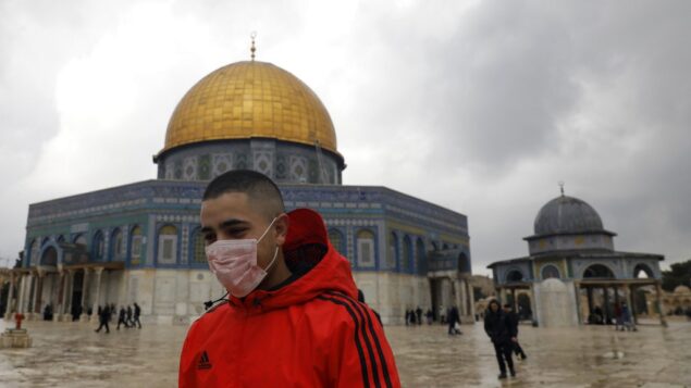 شاب فلسطيني يرتدي قناعا واقيا كإجراء حماية ضد فيروس كورنا COVID-19، يقف أمام قبة الصخرة في الحرم القدسي بالبلدة القديمة، 13 مارس، 2020. (Ahmad Gharabli/AFP)