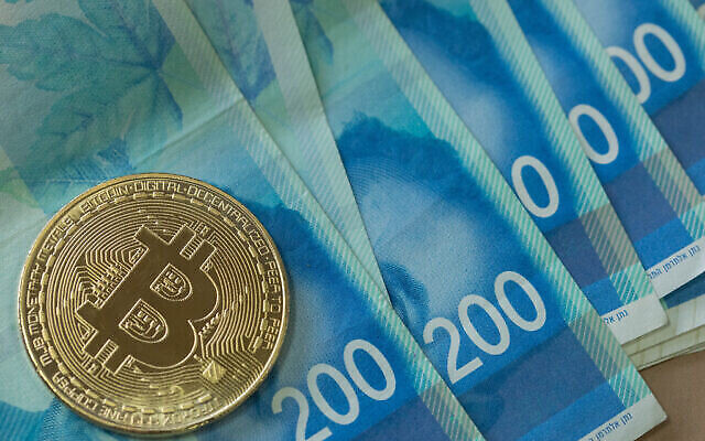 توضيحية: عملة معدنية تحمل شعار البيتكوين ، فوق عدة أوراق ورقية من الشيكل الإسرائيلي الجديد. (Noam Revkin Fenton/Flash90)