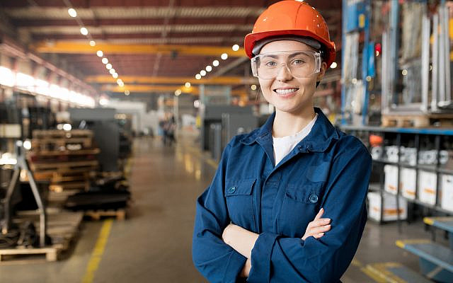 Young smiling female technician in blue uniform, protective eyeglasses and helmet working in modern factory