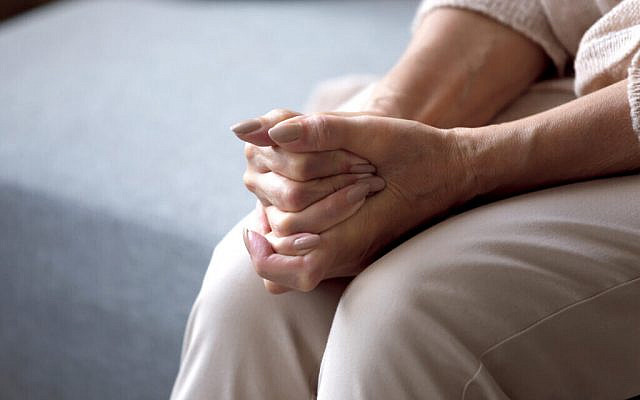 Elderly woman seated on couch holding hands together folded on lap feeling lonely (Via Jewish News)