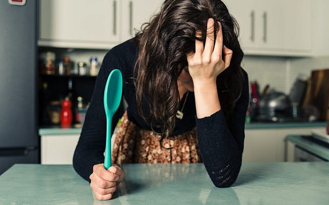 A sad young woman is standing in a kitchen with a spoon in her hand