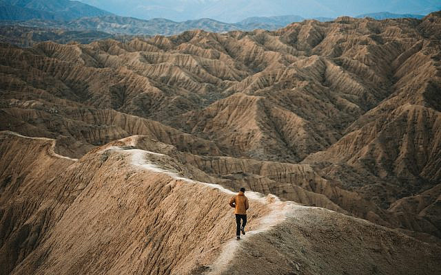 A lone figure advances along a narrow ridge across vast terrain, one step at a time, with no clear end in sight.