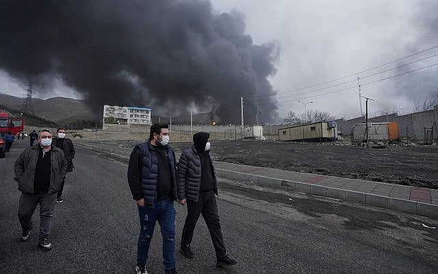 Four men walk as a thick plume of smoke from a US-Israeli strike on an oil storage facility late Saturday rises behind them in Tehran, Iran, Sunday, March 8, 2026. (AP Photo/Vahid Salemi)
