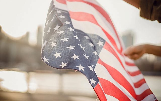 American flag waving in the city with blurred skyline background., as downloaded from Pexels (URL: https://www.pexels.com/photo/crop-person-holding-american-flag-in-wind-4560088/).