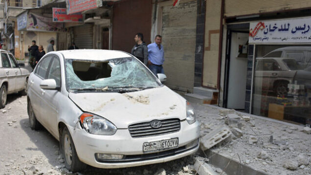 In this photo released by the Syrian official news agency SANA, people stand near a car that was damaged due to flying debris from a Syrian military base housing a weapons warehouse, in the country's central province of Homs, Syria, Friday, May 1, 2020. Explosions rocked a Syrian military base housing a weapons warehouse Friday in the country's central Homs province, and a local official said the cause was not immediately clear. (SANA via AP)