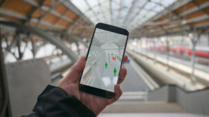 Older hand with a smartphone app for tracking the spread of infection which determines the contact persons of people who have been infected with the coronavirus, train station blurry in the background, copy space