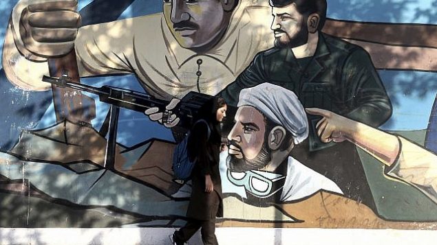 توضیح تصویر: یک زن ایرانی از مقابل دیوارنگاری از اعضای شبه نظامیان بسیجی، که ایرانیان را در مقابل دشمنان خود، متحد تصویر می کند، و بر دیوار یک بنای دولتی در میدان فلسطین در پایین شهر تهران نقاشی شده، می گذرد، تهران، ایران، 30 ژوئن 2018. (AP Photo/Ebrahim Noroozi)