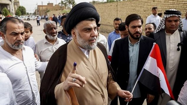 تصویر: مقتدی صدر، روحانی و رهبر شیعیان عراق، وسط-چپ، در میان جمعیتی که بیرون مرکز رأی گیری، در شهر مقدس نجف دور او حلقه زده اند، انگشت جوهری خود را نشان میدهد و پرچم ملی را در دست دارد، ۱۲ مه ۲۰۱۸. 
(Haidar Hamdani/AFP)