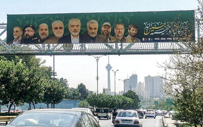 شرح عکس: اتوموبیلها در ۱۴ اوت ۲۰۲۴، از جاده-ای زیر پل عابر پیاده در تهران، که بانر بزرگی با تصویر قاسم سلیمانی فرمانده ترور-شدهٔ نیروی قدس سپاه پاسداران انقلاب اسلامی  (وسط، راست)، و چهره-های ترور-شدهٔ گروههای تحت حمایت ایران، نظیر اسمعیل هنیه از گروه تروریستی فلسطینی حماس (وسط)، ابو مهدی المهندس از حشد الشعبی عراق (وسط، چپ)، عماد مغنیه از حزب الله لبنان (سومی، راست) روی آن نصب است، میگذرند.
(Atta Kenare/AFP)