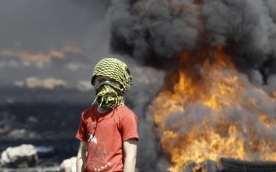 Un garçon palestinien masqué à côté de pneus en feu (Crédit : Jaafar Ashtiyeh/AFP)