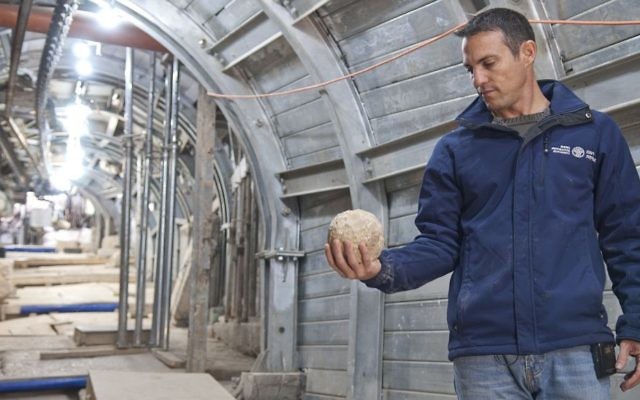 Nahshon Szanton tenant une pierre d'arbalète qui aurait été catapultée durant le siège de Jérusalem, dans une rue antique où des fouilles sont actuellement menées (Crédit : Shai Halevy/Autorisation : Autorité des antiquités israéliennes) Nahshon Szanton tenant une pierre d'arbalète qui aurait été catapultée durant le siège de Jérusalem, dans une rue antique où des fouilles sont actuellement menées (Crédit : Shai Halevy/Autorisation : Autorité des antiquités israéliennes)