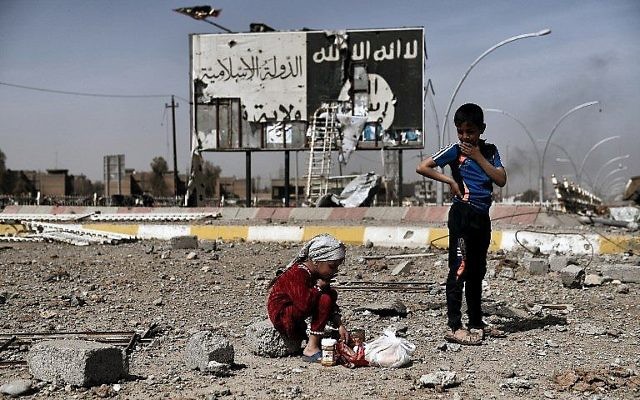 Cette photo prise le 12 mars 2017 montre des enfants irakiens assis dans les décombres d'une rue du quartier de Naplouse à Mossoul devant un tableau portant le logo de l'Etat islamique durant une offensive des forces de sécurité pour la reprise des quartiers ouest de la ville, aux mains de l'EI (Crédit : AFP PHOTO / ARIS MESSINIS)