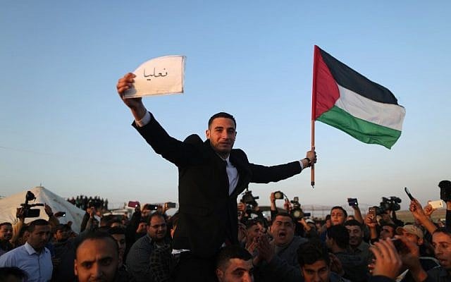 Les Palestiniens portent un jeune marié qui brandit le drapeau palestinien et tient une pancarte avec le nom du village de sa famille en IsraPel à la frontière de Gaza avec Israël (dans le fond), à l'est de Jabalia, le 29 mars 2018, avant la tenue d'un camp de protestation de six semaines (Crédit : AFP PHOTO / MAHMUD HAMS) Les Palestiniens portent un jeune marié qui brandit le drapeau palestinien et tient une pancarte avec le nom du village de sa famille en IsraPel à la frontière de Gaza avec Israël (dans le fond), à l'est de Jabalia, le 29 mars 2018, avant la tenue d'un camp de protestation de six semaines (Crédit : AFP PHOTO / MAHMUD HAMS)