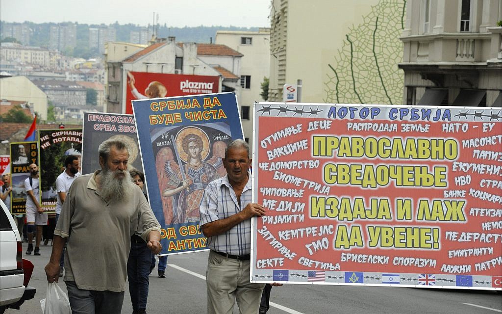 En juin 2018, des nationalistes serbes défilent dans une rue de Belgrade, portant des banderoles qui protestent contre la profanation des églises orthodoxes serbes et attribuent les problèmes économiques du pays à des "influences étrangères néfastes" telles que l'OTAN, la Banque mondiale, la télévision réalité, l'UE, les maçons, les aliments transgéniques, Israël et les États-Unis. (Larry Luxner/Times of Israel)