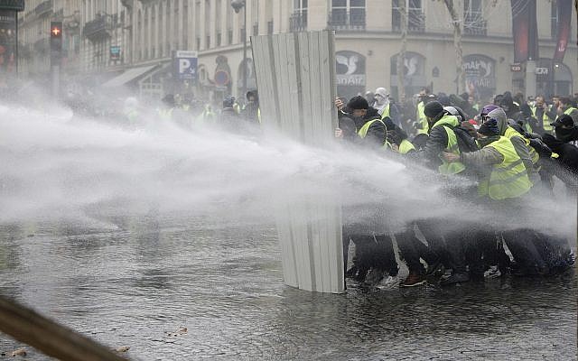 Les "Gilets jaunes" utilisent une barrière pour se protéger des canons à eau des policiers pendant une manifestation sur les Champs Elysées pour dénoncer la hausse des taxes sur les carburants,  le 24 novembre 2018 (Crédit : AP Photo/Kamil Zihnioglu)