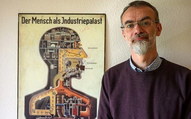 Le responsable de l'Institut d'anatomie de l'école de médecine du Brandenbourg, Andreas Winkelmann, pose près d'une affiche de 1926 détaillant le corps humain comme un "palais industriel", avril 2019. (Crédit : MACDOUGALL / AFP) Le responsable de l'Institut d'anatomie de l'école de médecine du Brandenbourg, Andreas Winkelmann, pose près d'une affiche de 1926 détaillant le corps humain comme un "palais industriel", avril 2019. (Crédit : MACDOUGALL / AFP)