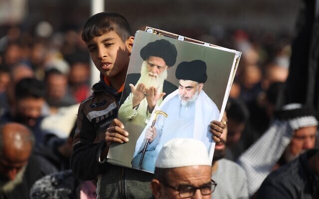 Un jeune garçon tient une affiche montrant les hauts responsables chiites l'Ayatollah Ali Sistan (à gauche) et Moqtada al-Sadr alors que les partisans de ce dernier participent aux prières du vendredi dans la banlieue de Bagdad, le 3 janvier 2020. (Crédit : AHMAD AL-RUBAYE / AFP)