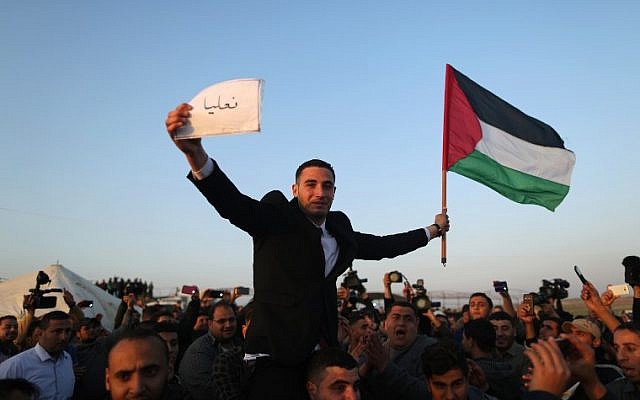 Les Palestiniens portent un jeune marié qui brandit le drapeau palestinien et tient une pancarte avec le nom du village de sa famille en IsraPel à la frontière de Gaza avec Israël (dans le fond), à l'est de Jabalia, le 29 mars 2018, avant la tenue d'un camp de protestation de six semaines (Crédit : AFP PHOTO / MAHMUD HAMS) Les Palestiniens portent un jeune marié qui brandit le drapeau palestinien et tient une pancarte avec le nom du village de sa famille en IsraPel à la frontière de Gaza avec Israël (dans le fond), à l'est de Jabalia, le 29 mars 2018, avant la tenue d'un camp de protestation de six semaines (Crédit : AFP PHOTO / MAHMUD HAMS)