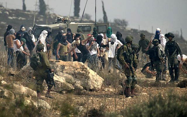 Photo d'illustration : Des soldats israéliens regardent des habitants d'implantation masqués jeter des pierres à des manifestants palestiniens (non-visibles sur la photo) pendant une manifestation contre la construction d'un avant-poste israélien aux abords du village de Turmusaya et de l'implantation de Shilo, au nord de Ramallah, en Cisjordanie, le 17 octobre 2019. (Crédit : JAAFAR ASHTIYEH / AFP) Photo d'illustration : Des soldats israéliens regardent des habitants d'implantation masqués jeter des pierres à des manifestants palestiniens (non-visibles sur la photo) pendant une manifestation contre la construction d'un avant-poste israélien aux abords du village de Turmusaya et de l'implantation de Shilo, au nord de Ramallah, en Cisjordanie, le 17 octobre 2019. (Crédit : JAAFAR ASHTIYEH / AFP)