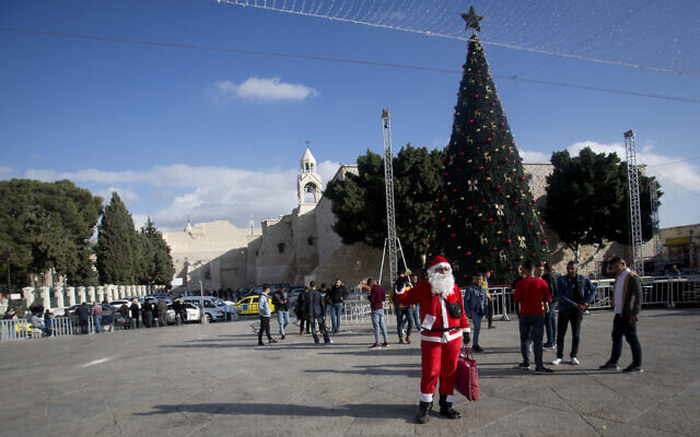 Un Palestinien portant un costume de père Noël accueille des visiteurs chrétiens à l'extérieur de l'église de la Nativité, traditionnellement considérée par les Chrétiens comme le lieu de naissance de Jésus-Christ, dans la ville cisjordanienne de Bethléem, le 5 décembre 2019. (Crédit : Majdi Mohammed / AP) Un Palestinien portant un costume de père Noël accueille des visiteurs chrétiens à l'extérieur de l'église de la Nativité, traditionnellement considérée par les Chrétiens comme le lieu de naissance de Jésus-Christ, dans la ville cisjordanienne de Bethléem, le 5 décembre 2019. (Crédit : Majdi Mohammed / AP)
