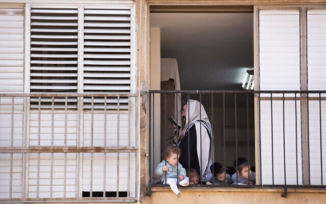 Un homme ultra-orthodoxe récite sa prière du matin dans son appartement alors que ses enfants regardent par la fenêtre lors d'un confinement imposé pour ralentir la propagation du coronavirus à Bnei Brak, le 8 avril 2020. (Crédit : AP Photo/Oded Balilty) Un homme ultra-orthodoxe récite sa prière du matin dans son appartement alors que ses enfants regardent par la fenêtre lors d'un confinement imposé pour ralentir la propagation du coronavirus à Bnei Brak, le 8 avril 2020. (Crédit : AP Photo/Oded Balilty)