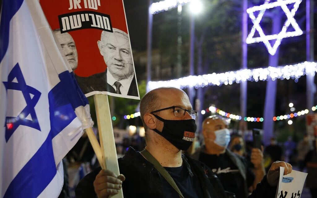 Un manifestant israélien portant un masque de protection avec le slogan "ministre du crime" en compagnie d'autres personnes respectant la distanciation de deux mètres imposée par le coronavirus brandit un panneau avec les visages du Premier ministre Benjamin Netanyahu et de Benny Gantz, ex-chef de Kakhol lavan, lors d'une manifestation sur la place Rabin de Tel Aviv, le 9 mai 2020 (Crédit : JACK GUEZ / AFP)