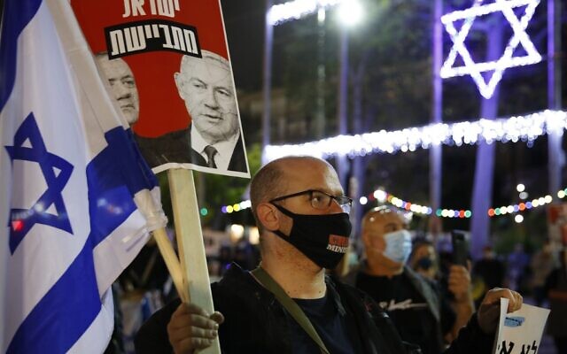 Un manifestant israélien portant un masque de protection avec le slogan "ministre du crime" en compagnie d'autres personnes respectant la distanciation de deux mètres imposée par le coronavirus brandit un panneau avec les visages du Premier ministre Benjamin Netanyahu et de Benny Gantz, ex-chef de Kakhol lavan, lors d'une manifestation sur la place Rabin de Tel Aviv, le 9 mai 2020 (Crédit : JACK GUEZ / AFP)