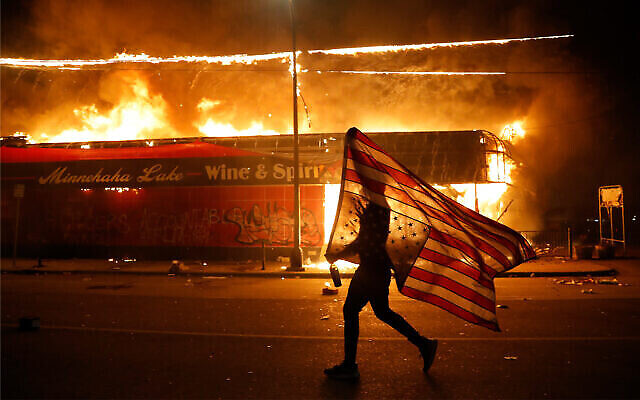 Un manifestant porte un drapeau américain à l'envers, signe de désespoir, à côté d'un immeuble en feu à Minneapolis, dans le Minnesota, après la mort de George Floyd, le 28 mai 2020. (Crédit : AP / Julio Cortez) Un manifestant porte un drapeau américain à l'envers, signe de désespoir, à côté d'un immeuble en feu à Minneapolis, dans le Minnesota, après la mort de George Floyd, le 28 mai 2020. (Crédit : AP / Julio Cortez)