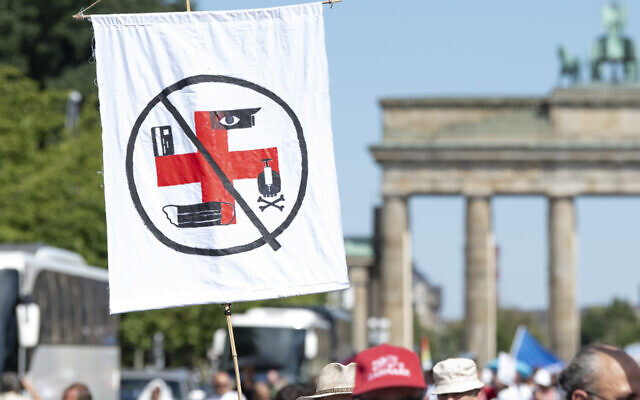 Le 1 août 2020 à Berlin, un drapeau sur lequel on peut voir différent symboles accrochés à une croix rouge former une croix gammée est tenu par un participant dans la manifestation contre les restrictions liées au coronavirus devant la Porte de Brandenburg. (Fabian Sommer/picture alliance via Getty Images via JTA) Le 1 août 2020 à Berlin, un drapeau sur lequel on peut voir différent symboles accrochés à une croix rouge former une croix gammée est tenu par un participant dans la manifestation contre les restrictions liées au coronavirus devant la Porte de Brandenburg. (Fabian Sommer/picture alliance via Getty Images via JTA)
