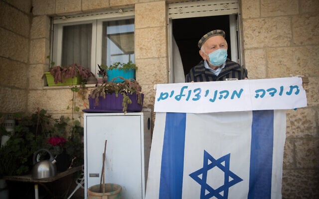Illustration : Joseph Kleinman, un survivant de la Shoah âgé de 90 ans, qui a survécu aux camps de la mort nazis d'Auschwitz et de Dachau, avec un masque et un drapeau israélien sous son porche à Jérusalem, pendant le jour du souvenir de l'Holocauste, le 21 avril 2020. (Crédit : Yonatan Sindel / Flash90)
