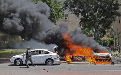 Un soldat israélien passe devant un véhicule en feu après qu'une roquette lancée depuis la bande de Gaza a frappé la ville d'Ashkelon, dans le sud d'Israël, le 11 mai 2021 (Crédit : JACK GUEZ / AFP)