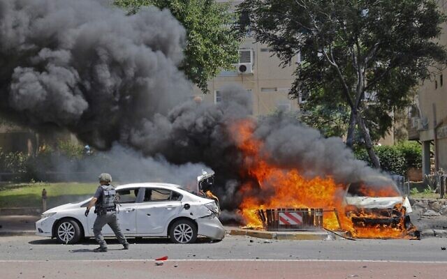 Un soldat israélien passe devant un véhicule en feu après qu'une roquette lancée depuis la bande de Gaza a frappé la ville d'Ashkelon, dans le sud d'Israël, le 11 mai 2021 (Crédit : JACK GUEZ / AFP)